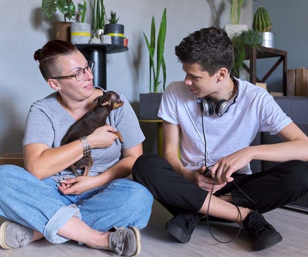 Happy talking mom and teenage son, parent and child sitting at home on the floor with pet little dog toy terrier, real friendly loving family